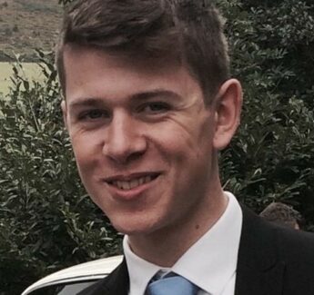 Headshot oung man in black jacket, white shirt and pale blue tie looking to his right, with tree in background