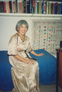 Professor Hisako Ikeda-Wolstencroft sat on her bed in her office, the bed has a blue cover. Professor Hisako Ikeda-Wolstencroft is smiling into the camera wearing a satin long dress. There is a book shelf full of books above the bed and a wall hanging underneath the bookshelf.