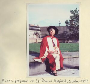 Professor Hisako Ikeda-Wolstencroft sat on a wall outside at St. Thomas Hospital in the sun, in her red and white graduation gown with a black tudor bonet.