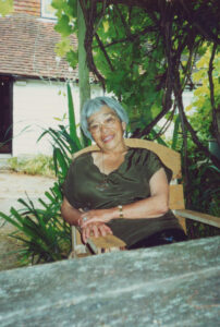 Professor Hisako Ikeda-Wolstencroft sat outside on a wooden chair behind a wooden table smiling at the camera. There are green plants and vines behind Professor Hisako Ikeda-Wolstencroft and a building in the background.